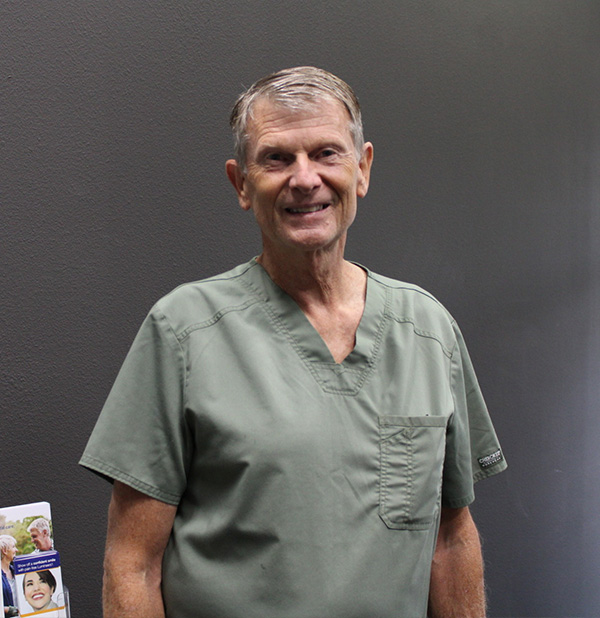 The image shows an older man wearing a light green scrub top, standing indoors with a professional background that appears to be a medical office.