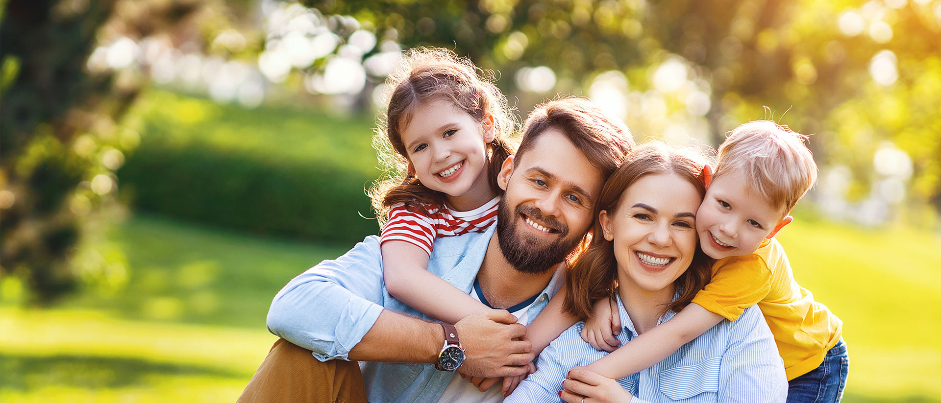 A family of four posing for a photo in an outdoor setting, with the father holding two children and the mother standing behind them.