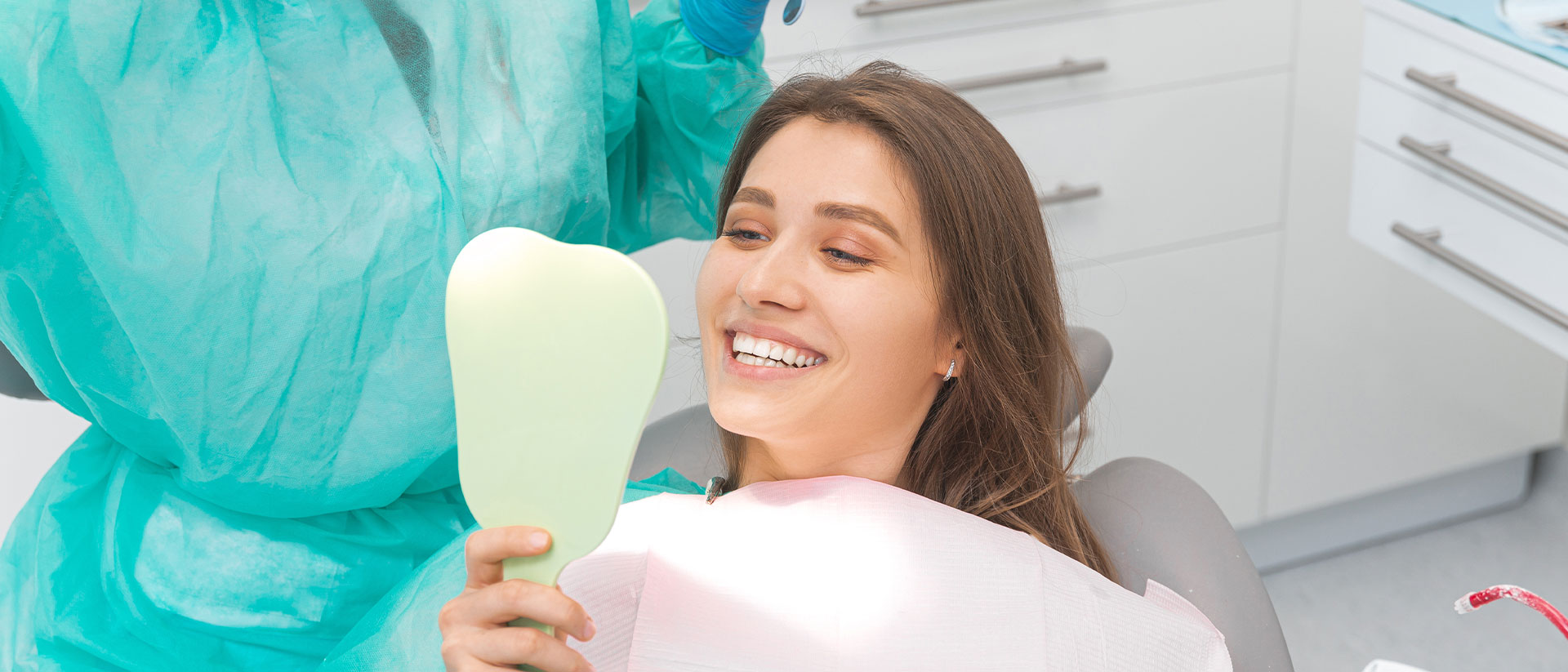 A woman in a dental chair is smiling at the camera while holding a toothbrush, with a dental professional in protective clothing standing behind her.