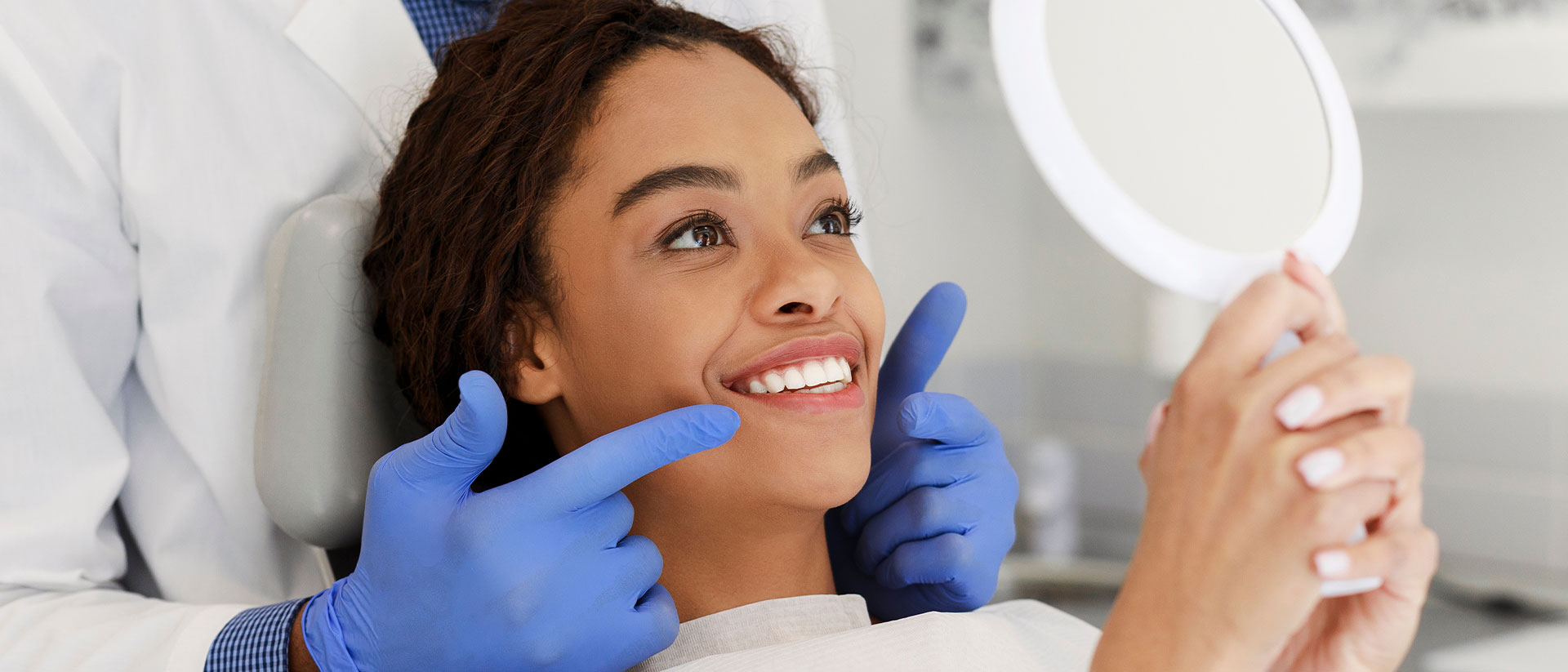 A woman with a wide smile is seated in a dental chair, receiving dental care from a professional who is holding a mirror.