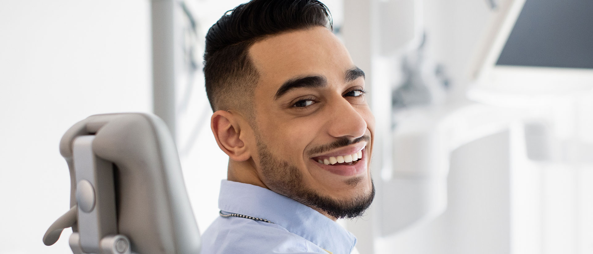 The image is a photograph of a smiling man in a dental office, sitting in a chair with his head tilted slightly to the side, wearing a blue shirt and a necklace, looking directly at the camera.