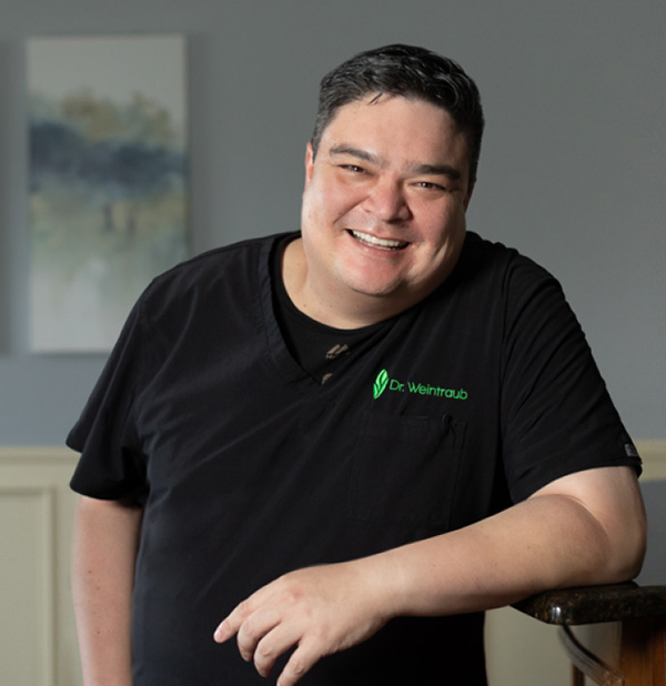 A man in a black shirt and a logo on the left chest area is smiling, leaning against a wall with a whiteboard behind him.