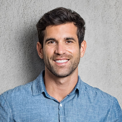 A smiling man with short hair, wearing a light blue shirt and standing against a gray background.