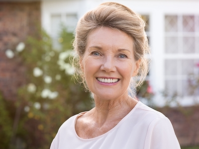 Smiling older woman with blonde hair, wearing a white top, standing in front of a house with a brick wall and green plants.