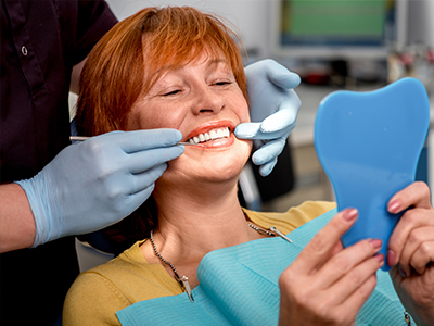 The image shows a woman sitting in a dental chair, receiving dental care, with a dental mirror and a toothbrush in her mouth.