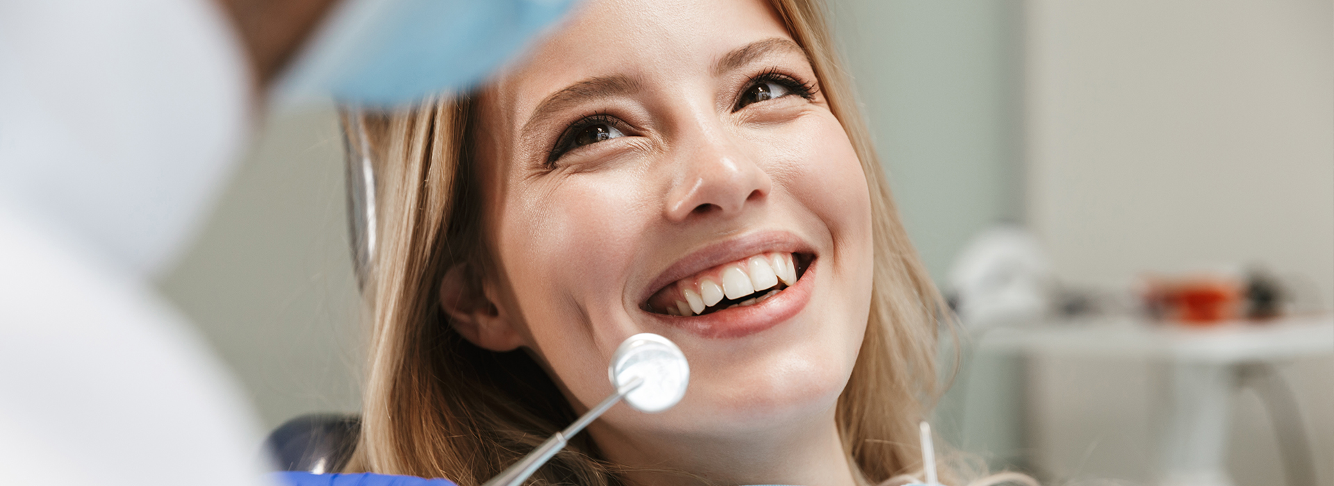 The image shows a woman with a smile, sitting in a dental chair, while a dentist is performing a procedure on her teeth.