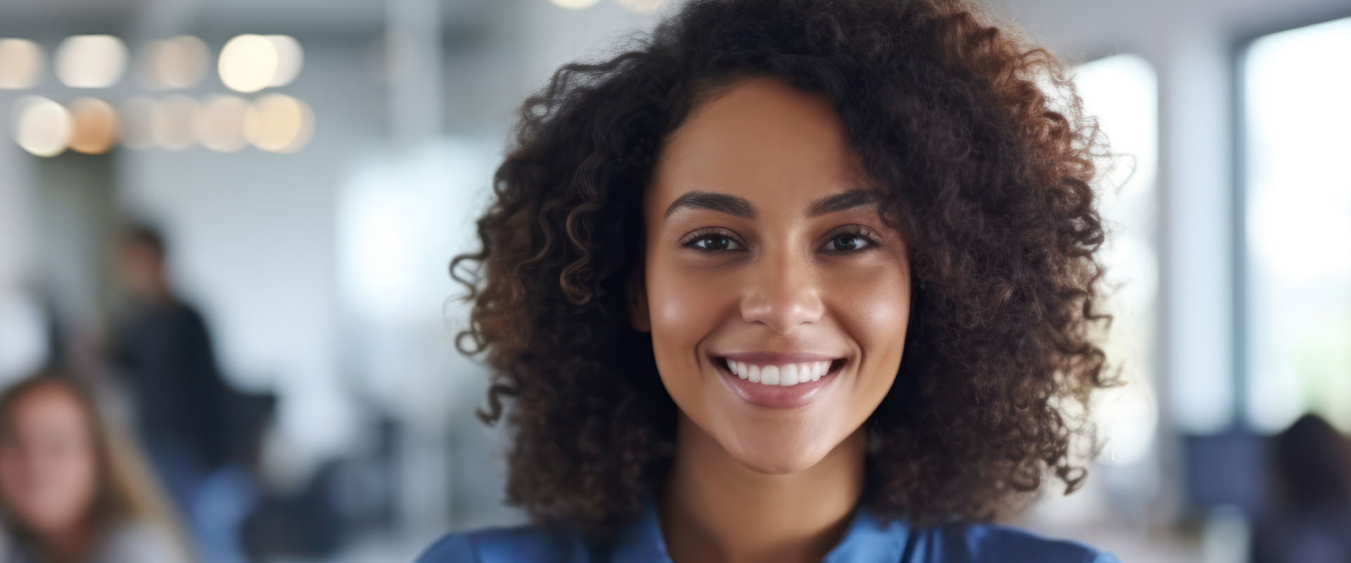 The image shows a person, likely a woman based on the visible facial features, smiling at the camera. She has dark hair and appears to be in an indoor setting with a blurred background that suggests an office environment.