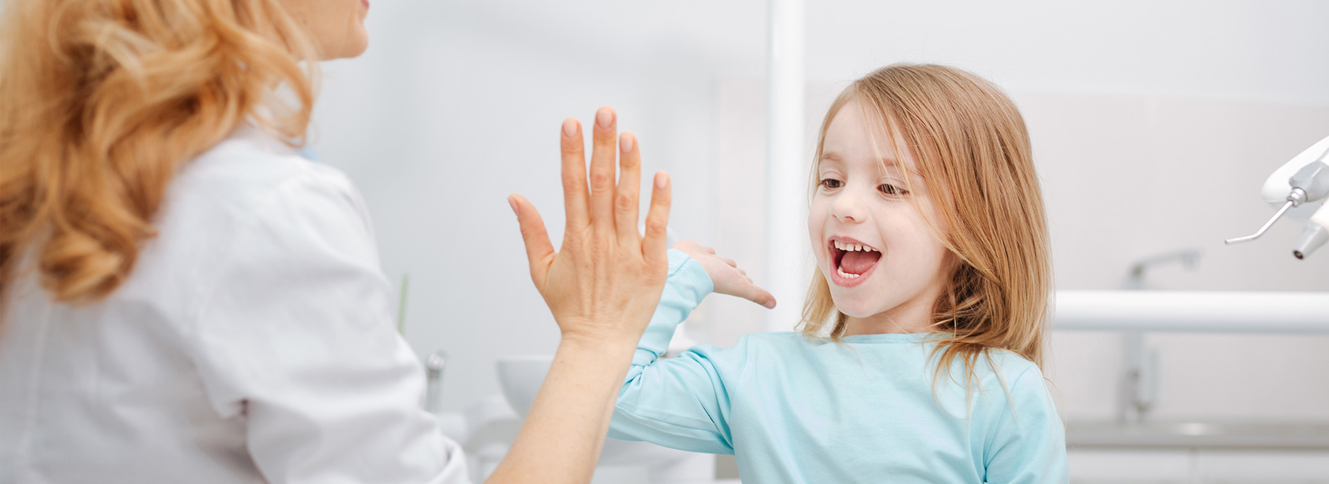 A young girl in a blue shirt is smiling and waving at the camera, with an adult woman s hand visible in the foreground.