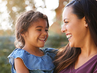 The image is a photograph of a woman and a child, both smiling and looking at the camera. The woman appears to be an adult with long hair and is wearing a dark top, while the child has short hair and is dressed in a light-colored outfit with a denim bib. They are outdoors during daylight, as indicated by natural lighting.