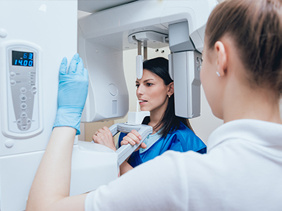 A woman in a blue uniform stands next to a modern medical imaging machine, with another individual observing the equipment.