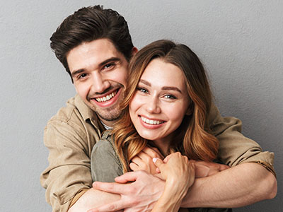 A young couple embracing and smiling, with the man wearing a short-sleeved shirt and the woman in a long-sleeve top.