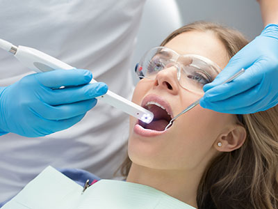 A woman in a dental chair receiving oral care with a dental device and a professional in protective attire assisting.