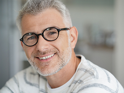 The image shows a man with glasses, smiling at the camera. He has short hair and is wearing a white shirt. The background is blurred but appears to be an indoor setting with natural light.