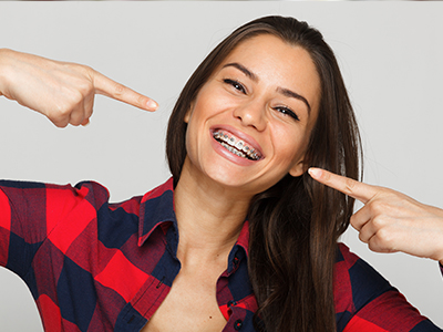 A woman with a gap-toothed smile pointing to her teeth, wearing a plaid shirt and standing against a white background.