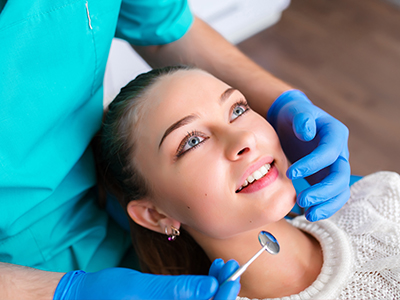 The image shows a woman sitting in a dental chair, smiling at the camera, with a dentist standing behind her, performing a dental procedure.