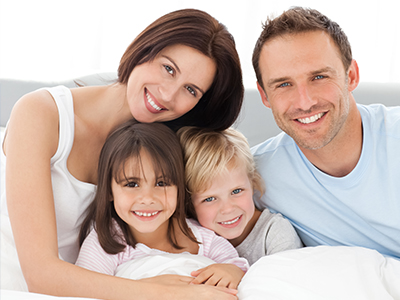 A family of four, including two adults and two children, smiling together on a bed with a white background.