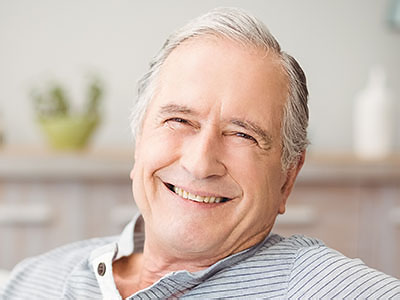 An elderly man with white hair, wearing a blue shirt and a smile on his face, is sitting in a chair.