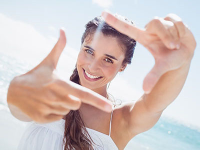 The image shows a woman outdoors, smiling and holding up her hand with fingers spread to make a frame around the camera. She is wearing sunglasses and has long hair.