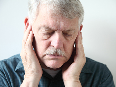 The image shows an older man with a beard and mustache, holding his hand to his ear as if listening intently or experiencing discomfort. He has a serious expression on his face.